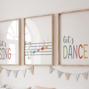 May include: Three framed art prints with wooden frames. The prints feature the phrases "let's SING" and "let's DANCE" in colourful letters, and a musical staff with notes. A banner of small flags hangs below.