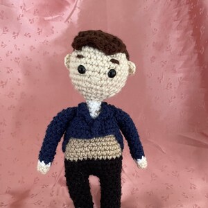 May include: A crocheted doll wearing a blue jacket, brown pants, and a white shirt. The doll has brown hair, a beige face, and black eyes.