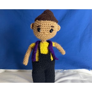 May include: A crocheted doll with brown hair, a yellow shirt, black overalls, and a purple scarf. The doll has black eyes and a neutral expression.