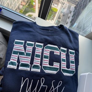 Puede incluir: Sudadera azul marino con las letras "NICU" en un diseño a rayas verdes, blancas y granates. Debajo de las letras, la palabra "nurse" está escrita en cursiva. La sudadera está doblada sobre un alféizar.