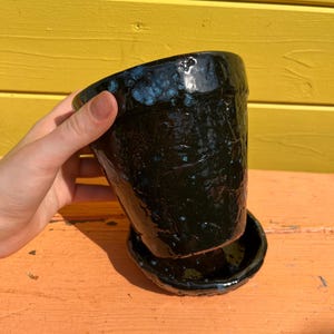 Puede incluir: Una maceta de cerámica negra con un platillo a juego. La maceta tiene un acabado brillante con motas azules. La maceta está ligeramente inclinada, revelando su interior. El platillo es redondo y tiene un patrón moteado similar.