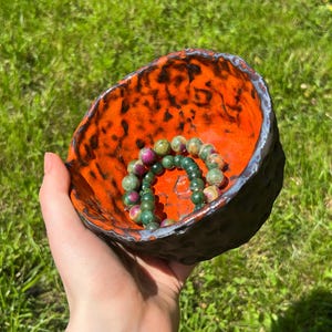 Op de afbeelding: Een handgemaakte keramische kom met een getextureerde, donkere buitenkant en een feloranje binnenkant. De kom bevat twee kralenarmbanden met groene en roze stenen. De kom wordt in een hand gehouden tegen een achtergrond van groen gras.