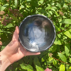 Może przedstawiać: Mała, ciemnoszara ceramiczna miska z falistym brzegiem. Misa ma błyszczącą, odbijającą światło powierzchnię. Tło przedstawia zielone liście i różowe kwiaty. Misa jest trzymana w dłoni.