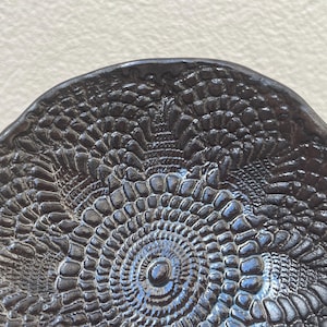 Puede incluir: Un plato de cerámica oscura y texturizada con un patrón detallado en relieve. La superficie del plato tiene una calidad brillante y reflectante, con toques de azul y negro. El diseño recuerda al encaje.