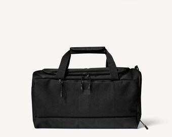 May include: Black duffel bag with a rectangular shape. The bag has two handles on top and a zippered compartment on the front. Additional zippers are visible on the sides. The bag is made of a durable, textured material.