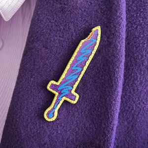 May include: A purple sword-shaped enamel pin with a yellow outline and blue zigzag design. The pin is attached to a purple blazer, with a white shirt and button-up collar visible in the background.