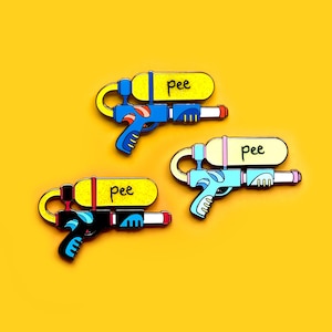 May include: Three enamel pins shaped like water guns with the word "pee" written on them. The water guns are blue, yellow, and pink.