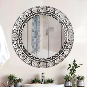 Puede incluir: Un espejo decorativo redondo con un marco plateado y negro adornado. El marco presenta un diseño floral y de volutas detallado. El espejo está colgado en una pared blanca sobre un lavabo de baño con plantas.