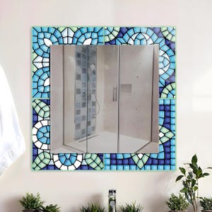 May include: Square mirror with a mosaic tile border in shades of blue, green, and white. The mosaic design features a repeating pattern of curved shapes and geometric tiles. The mirror reflects a shower stall.