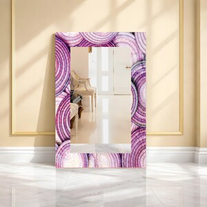May include: A rectangular mirror with a decorative frame featuring a repeating pattern of purple and white concentric circles. The mirror is leaning against a wall, reflecting a room with a chair and a door.