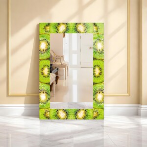 May include: A rectangular mirror with a frame decorated with green and yellow kiwi fruit slices. The mirror reflects a room with a white chair and door. The frame is made of a repeating pattern of kiwi fruit.