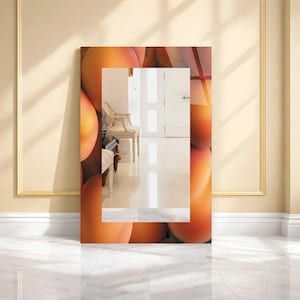 May include: A rectangular mirror leans against a wall, featuring a vibrant peach-themed design. The mirror's frame is adorned with a repeating pattern of large, ripe peaches in shades of orange and yellow. The mirror reflects a room with a white chair and door.