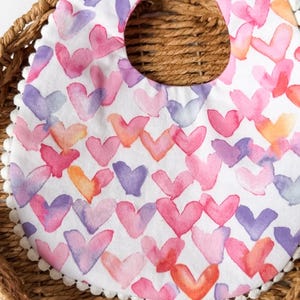May include: A white baby bib with a watercolor heart pattern in shades of pink, purple, and orange. The bib has a white pom-pom trim and a white snap closure.