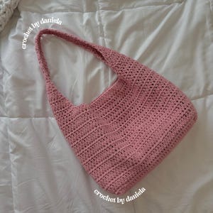 Peut inclure: Un sac &agrave; bandouli&egrave;re en crochet rose poudr&eacute; avec une longue sangle. Le sac est fait &agrave; la main avec un motif de point textur&eacute;. Les mots "crochet by daniela" sont &eacute;crits en cursive sur le sac. Le sac est sur un fond blanc.