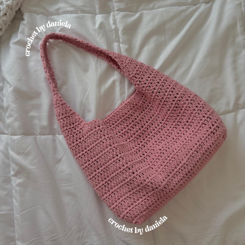 Peut inclure: Un sac &agrave; bandouli&egrave;re en crochet rose poudr&eacute; avec une longue sangle. Le sac est fait &agrave; la main avec un motif de point textur&eacute;. Les mots "crochet by daniela" sont &eacute;crits en cursive sur le sac. Le sac est sur un fond blanc.