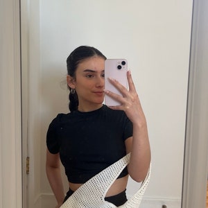 Puede incluir: Una mujer con un top negro corto y pantalones cortos negros sostiene una bolsa de mano de crochet blanca. Se est&aacute; tomando una selfie en un espejo.