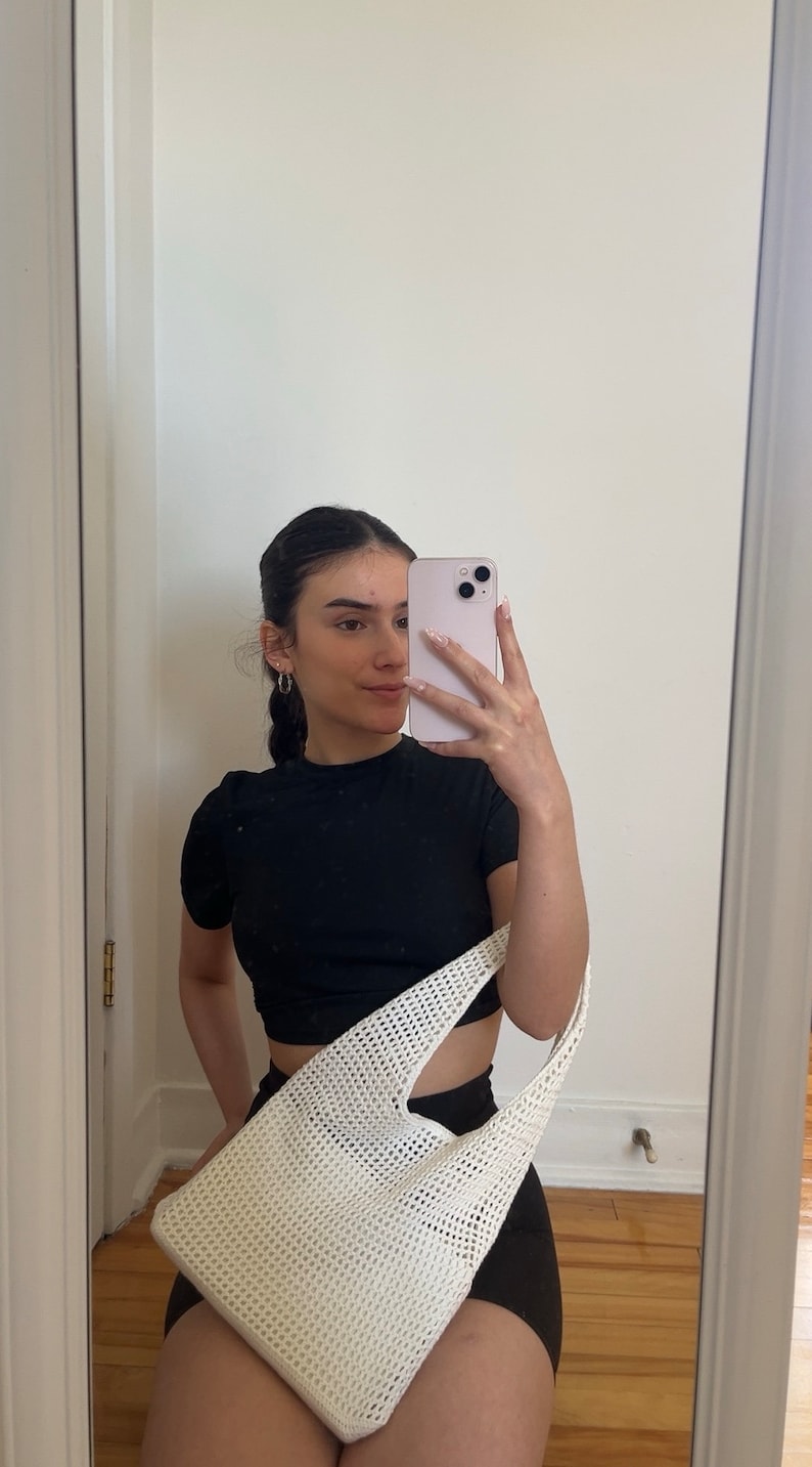 Puede incluir: Una mujer con un top negro corto y pantalones cortos negros sostiene una bolsa de mano de crochet blanca. Se est&aacute; tomando una selfie en un espejo.