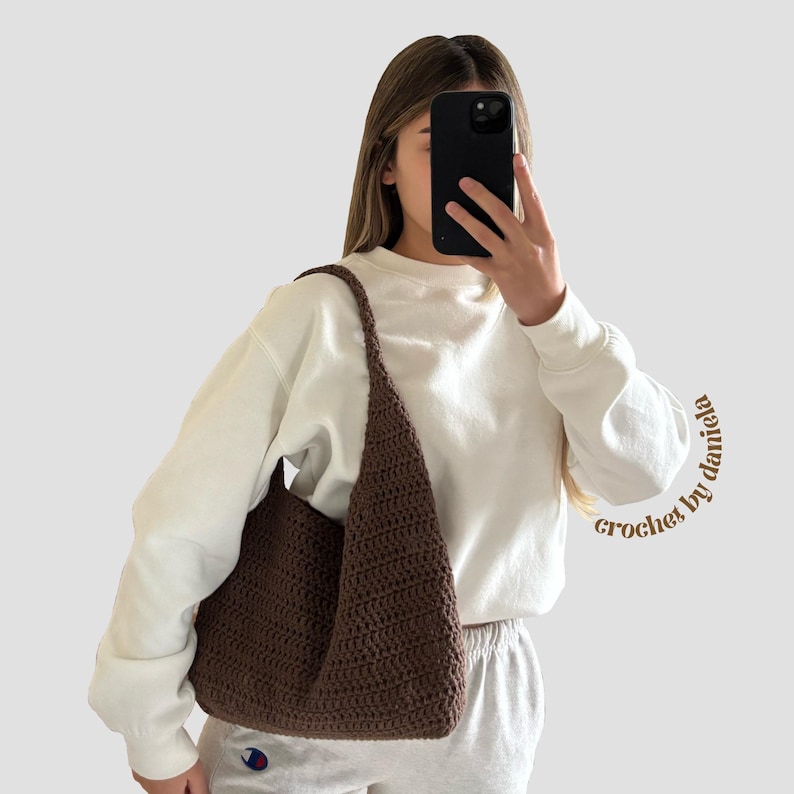 Peut inclure: Un sac fourre-tout en crochet marron avec une longue bandouli&egrave;re. Le sac est de couleur unie et de forme triangulaire. Les mots "crochet by daniela" sont &eacute;crits en cursive sur le c&ocirc;t&eacute; droit de l'image.