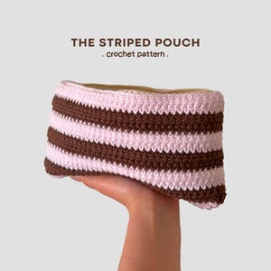 May include: A rectangular crochet pouch featuring horizontal stripes in alternating light pink and dark brown yarn. The text THE STRIPED POUCH . crochet pattern . is printed in dark brown above the pouch against a plain light background.