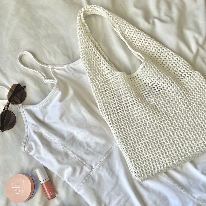 Puede incluir: Una bolsa de mano de crochet blanca con un top blanco, gafas de sol y maquillaje sobre una superficie blanca.