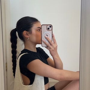 Puede incluir: Un bolso tote de crochet blanco con una correa negra. El bolso lo lleva una persona que lleva una camiseta negra y pantalones cortos negros.
