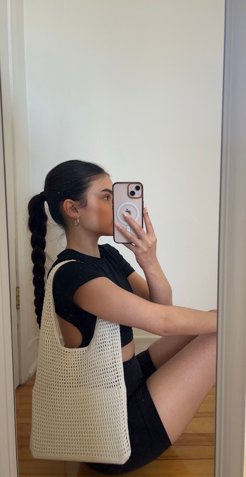 Puede incluir: Un bolso tote de crochet blanco con una correa negra. El bolso lo lleva una persona que lleva una camiseta negra y pantalones cortos negros.