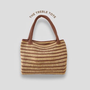 Könnte beinhalten: Gehäkelte Tragetasche mit horizontalen Streifen in Hellbraun und Dunkelbraun. Die Tasche hat zwei passende braune Griffe und den Text "THE TREBLE TOTE" über der Tasche.