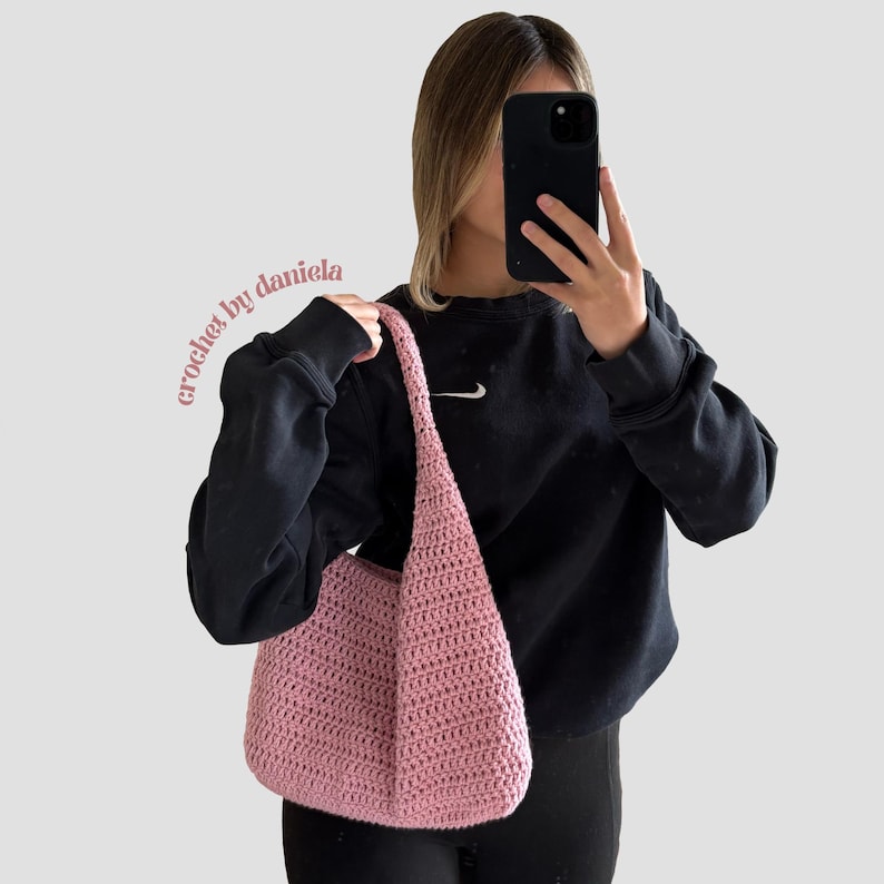 Peut inclure: Un sac &agrave; bandouli&egrave;re rose clair, crochet&eacute;, avec une longue sangle. Le sac a un fond arrondi et un motif de point textur&eacute;. Les mots "crochet by daniela" sont &eacute;crits en rose sur le c&ocirc;t&eacute; gauche de l'image.
