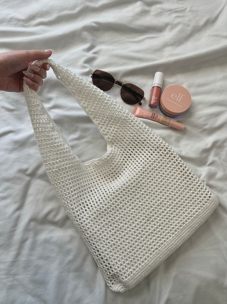 Puede incluir: Bolso tote de malla de crochet blanco con una sola correa. El bolso est&aacute; sobre una superficie blanca con gafas de sol, brillo de labios y productos de maquillaje.