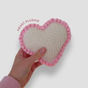 May include: A heart-shaped plushie with a cream-colored center and a pink crocheted border. The text "HEART PLUSHIE" is printed on the upper left side. The plushie is held in a hand.