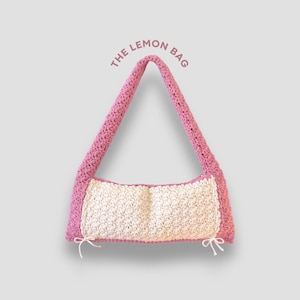May include: A handmade crochet bag with a pink strap and trim, and a cream-colored center. The text "THE LEMON BAG" is arched above the bag. White ties are attached to the bottom corners of the bag.