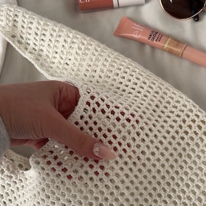 Puede incluir: Una bolsa de mano de crochet blanca sobre un fondo blanco. La bolsa est&aacute; hecha de un material de malla fina. Hay varios productos de maquillaje en el fondo, incluyendo un compacto rosa con el texto "elf.  Mixed Glow Setting Powder", un tubo de brillo de labios rosa con el texto "elf", y un tubo de iluminador rosa con el texto "elf. Halo Glow Beauty Wand.  Highlighter".