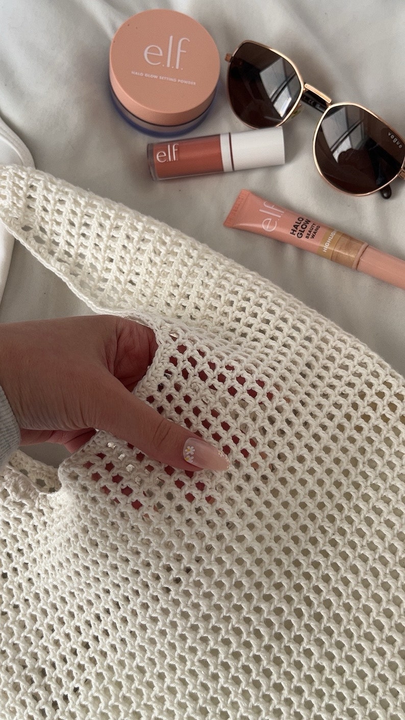 Puede incluir: Una bolsa de mano de crochet blanca sobre un fondo blanco. La bolsa est&aacute; hecha de un material de malla fina. Hay varios productos de maquillaje en el fondo, incluyendo un compacto rosa con el texto "elf.  Mixed Glow Setting Powder", un tubo de brillo de labios rosa con el texto "elf", y un tubo de iluminador rosa con el texto "elf. Halo Glow Beauty Wand.  Highlighter".