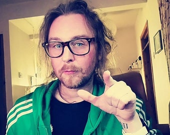 Op de afbeelding: Een persoon met een zwart montuur bril, een groene jas met witte strepen en een zwart t-shirt. De persoon heeft een baard en een polsband. Een tatoeage met Romeinse cijfers is zichtbaar op de pols.