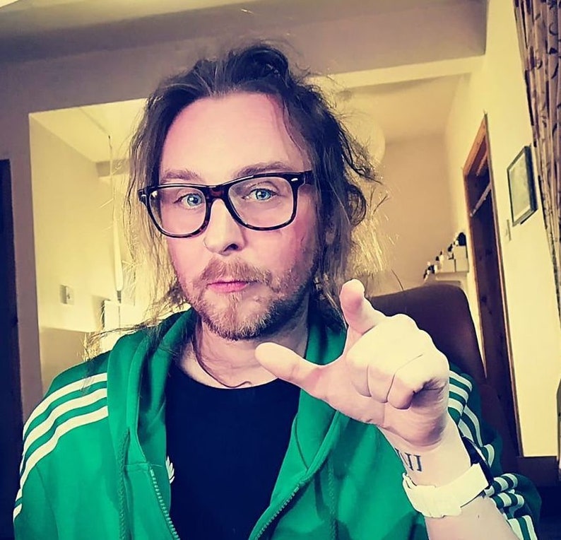 Pode incluir: Uma pessoa usando &oacute;culos com arma&ccedil;&atilde;o preta, uma jaqueta verde com z&iacute;per e listras brancas e uma camiseta preta. A pessoa tem barba e uma pulseira. Uma tatuagem com algarismos romanos &eacute; vis&iacute;vel no pulso.