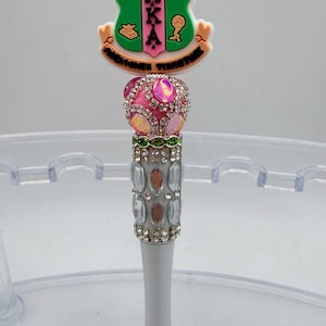 May include: A decorative pen featuring a green and pink emblem with the letters "AKA". The pen has a pink jeweled top, a silver barrel adorned with rhinestones, and a silver tip. The pen is designed for writing.