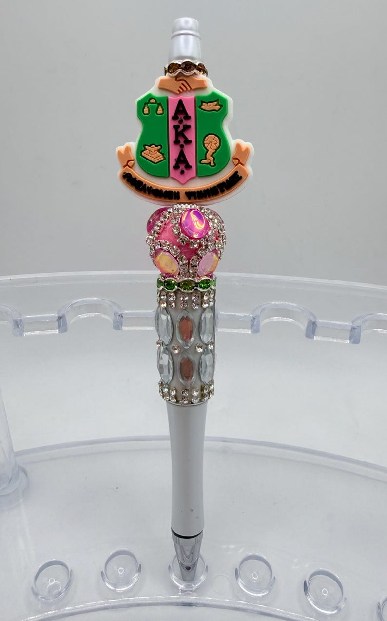 May include: A decorative pen featuring a green and pink emblem with the letters "AKA". The pen has a pink jeweled top, a silver barrel adorned with rhinestones, and a silver tip. The pen is designed for writing.