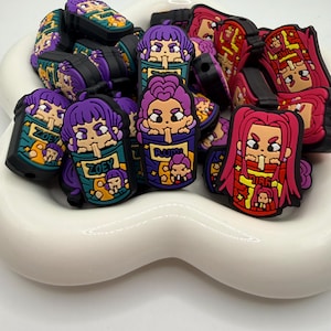 May include: A collection of colorful cartoon character charms in various designs. The charms feature characters with purple, red, and yellow hair, and are set against a white background. Some charms have the text "ZOE" and "RUMI".