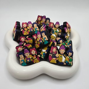 May include: A collection of colorful cartoon character shoe charms in a white, flower-shaped dish. The charms feature women with various hair colors and outfits, holding drinks. The charms are black, yellow, pink, purple, and green.