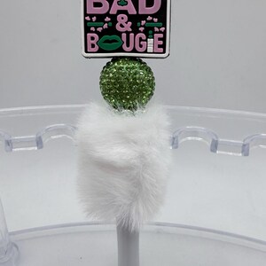 May include: A white pen with a fluffy white base, a green rhinestone ball, and a rectangular topper. The topper has the words "BAD & BOUJEE" in pink and green, and a silver crown. The pen is in a clear holder.