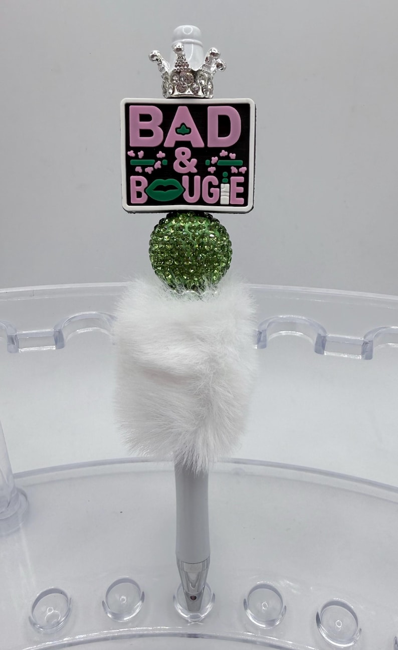 May include: A white pen with a fluffy white base, a green rhinestone ball, and a rectangular topper. The topper has the words "BAD & BOUJEE" in pink and green, and a silver crown. The pen is in a clear holder.
