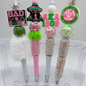 May include: A collection of decorative pens with various embellishments. The pens feature designs such as crowns, text that reads "BAD & BOUGIE", and the letters "AKA 1908". The pens are adorned with rhinestones, pearls, and fluffy accents.