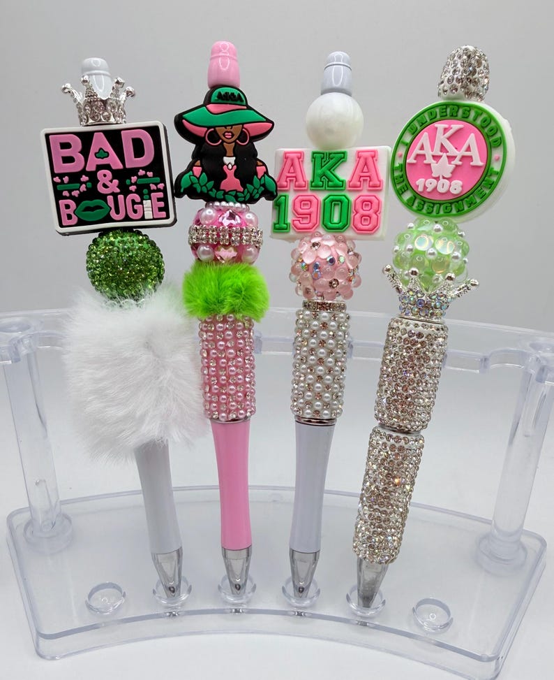 May include: A collection of decorative pens with various embellishments. The pens feature designs such as crowns, text that reads "BAD & BOUGIE", and the letters "AKA 1908". The pens are adorned with rhinestones, pearls, and fluffy accents.