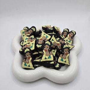 May include: A collection of black, yellow, and teal charms in a white, flower-shaped dish. The charms feature a cartoon woman with a tiara, necklace, and gloves. The charms are likely for jewelry making or other crafts.