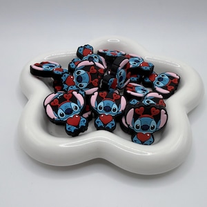 May include: A collection of blue, black, and pink Stitch character charms, each holding a red heart. The charms are arranged in a white, flower-shaped dish. The charms have a cartoon design.