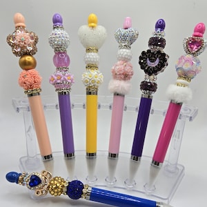 May include: A collection of decorative pens in various colors, including peach, purple, yellow, pink, and fuchsia. Each pen features a unique beaded design on top, with embellishments like flowers, hearts, and crystals. One blue pen with a jeweled top rests in front.