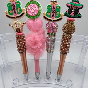 May include: Four decorative pens with various toppers, including a crown, a heart, and a woman in a hat. The pens have rose gold, silver, and pink barrels, and are displayed in a clear plastic holder. The toppers feature the letters AKA and the year 1908.