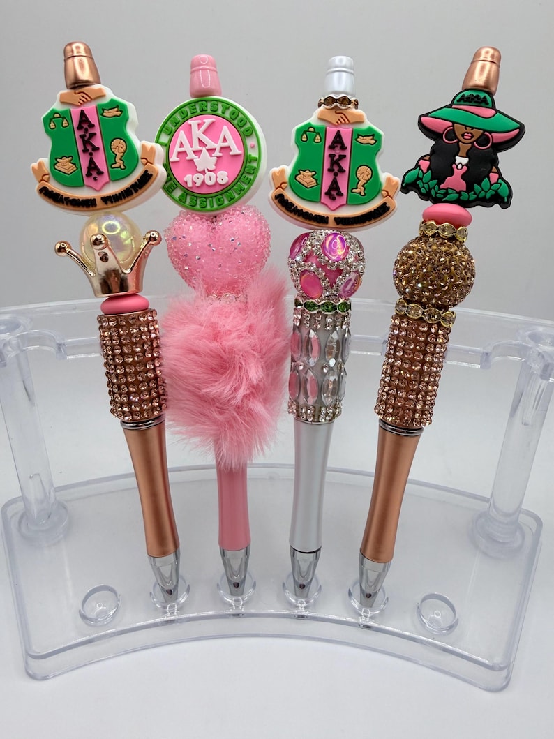 May include: Four decorative pens with various toppers, including a crown, a heart, and a woman in a hat. The pens have rose gold, silver, and pink barrels, and are displayed in a clear plastic holder. The toppers feature the letters AKA and the year 1908.