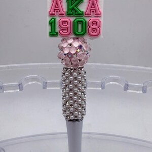 May include: A decorative pen with a white barrel and a beaded handle. The pen features a pink and green "AKA 1908" design, a pink crystal ball, and a white top.