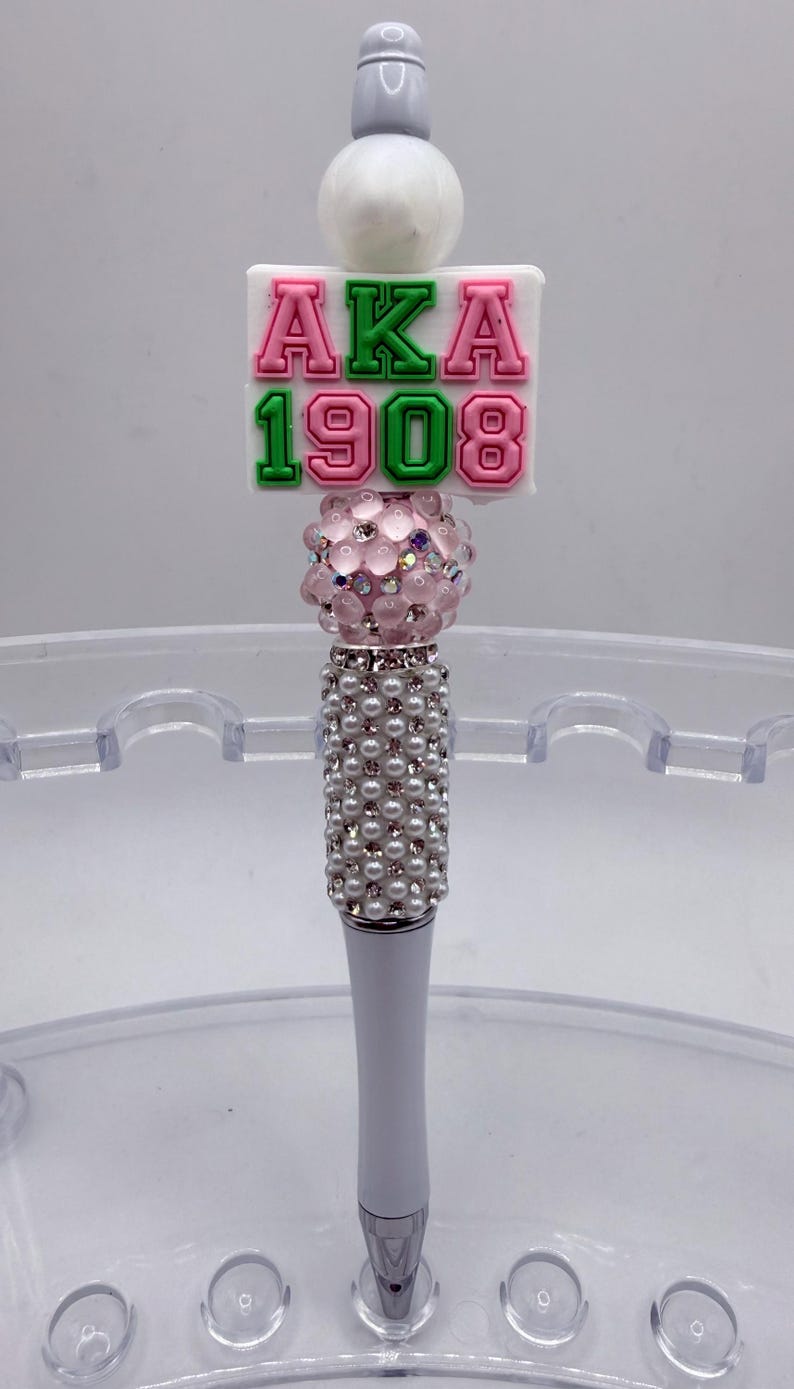 May include: A decorative pen with a white barrel and a beaded handle. The pen features a pink and green "AKA 1908" design, a pink crystal ball, and a white top.
